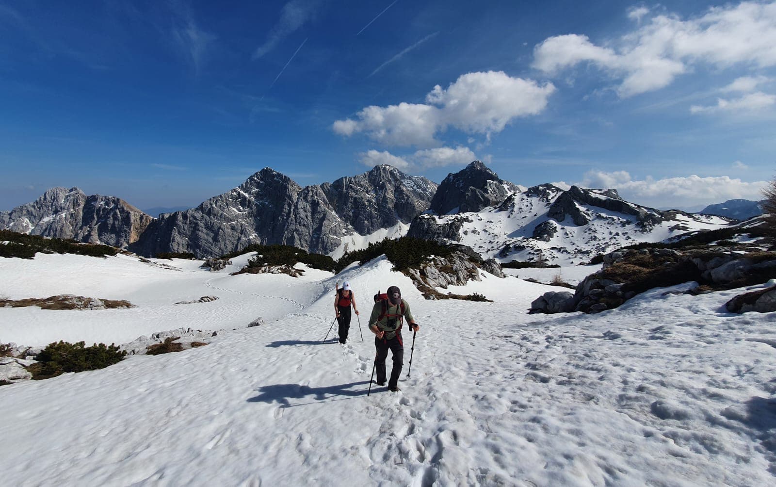Hikers trekking through snow toward jagged, snow-capped mountains under a blue sky near Kredarica.