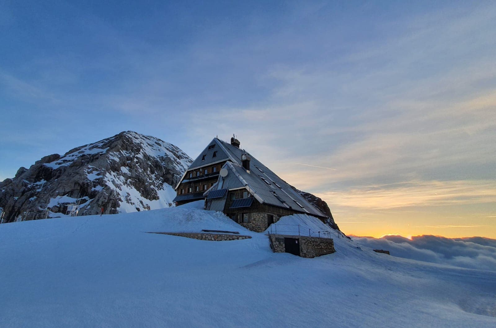 Kredarica mountain hut in deep snow near a rocky peak at winter sunset above clouds.