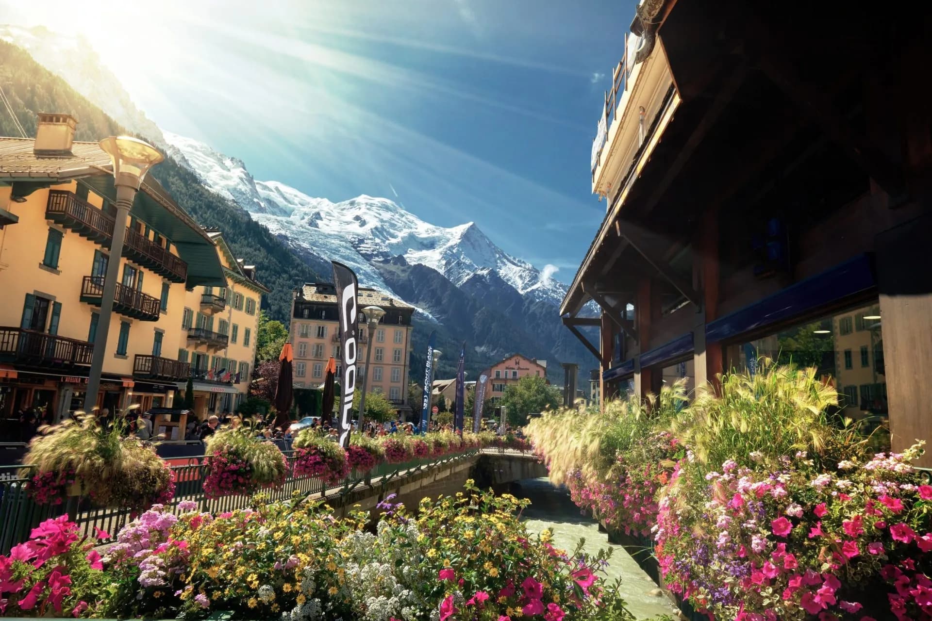Alpine town street with colorful flowers, river, and snow-capped mountains under bright sun.