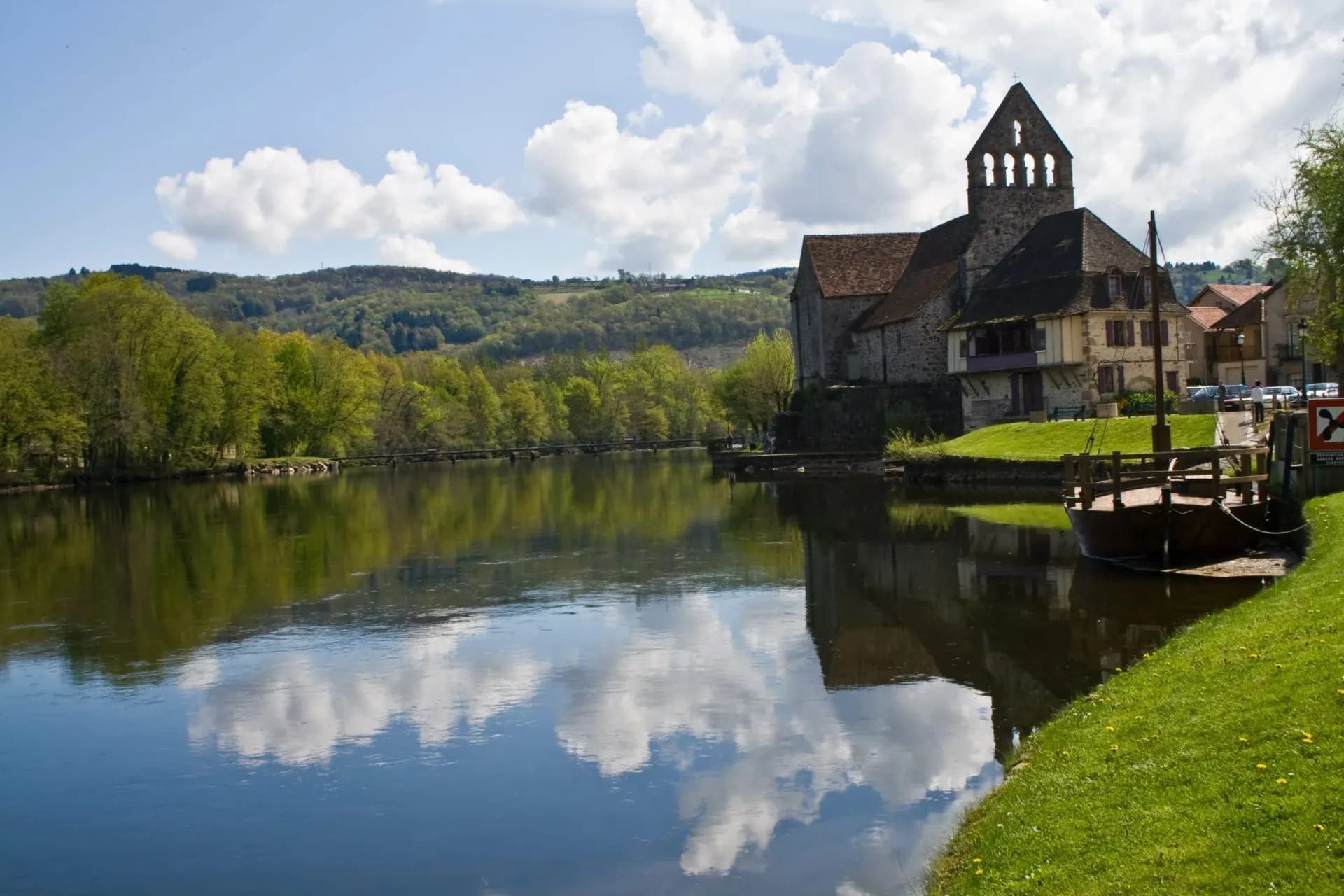 Stone church and boat moored on riverbank in Beaulieu-sur-Dordogne with reflections.