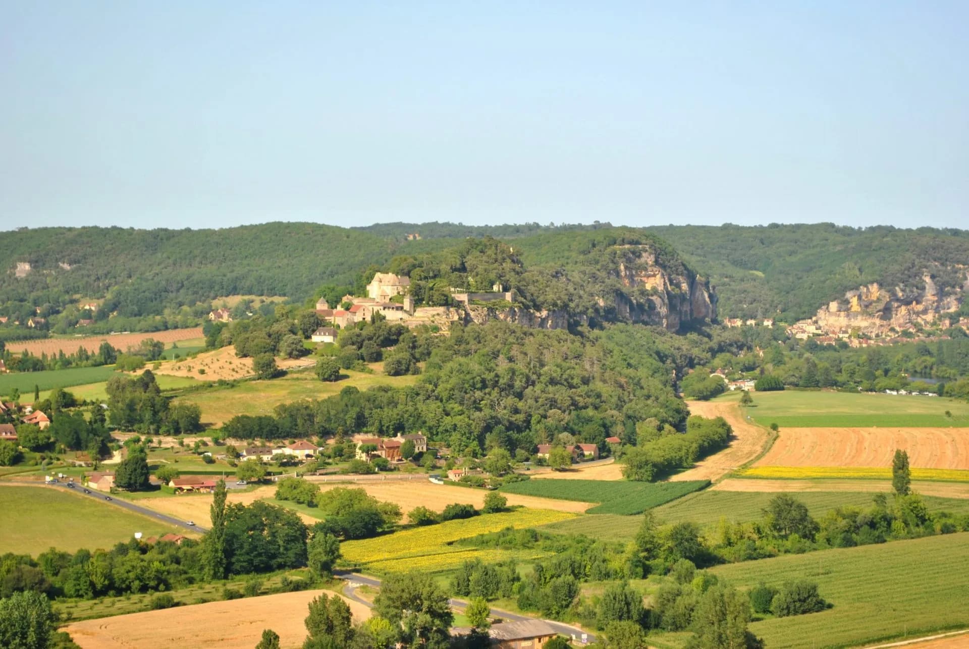 Village atop a wooded cliff overlooking green and yellow fields near Loubressac.