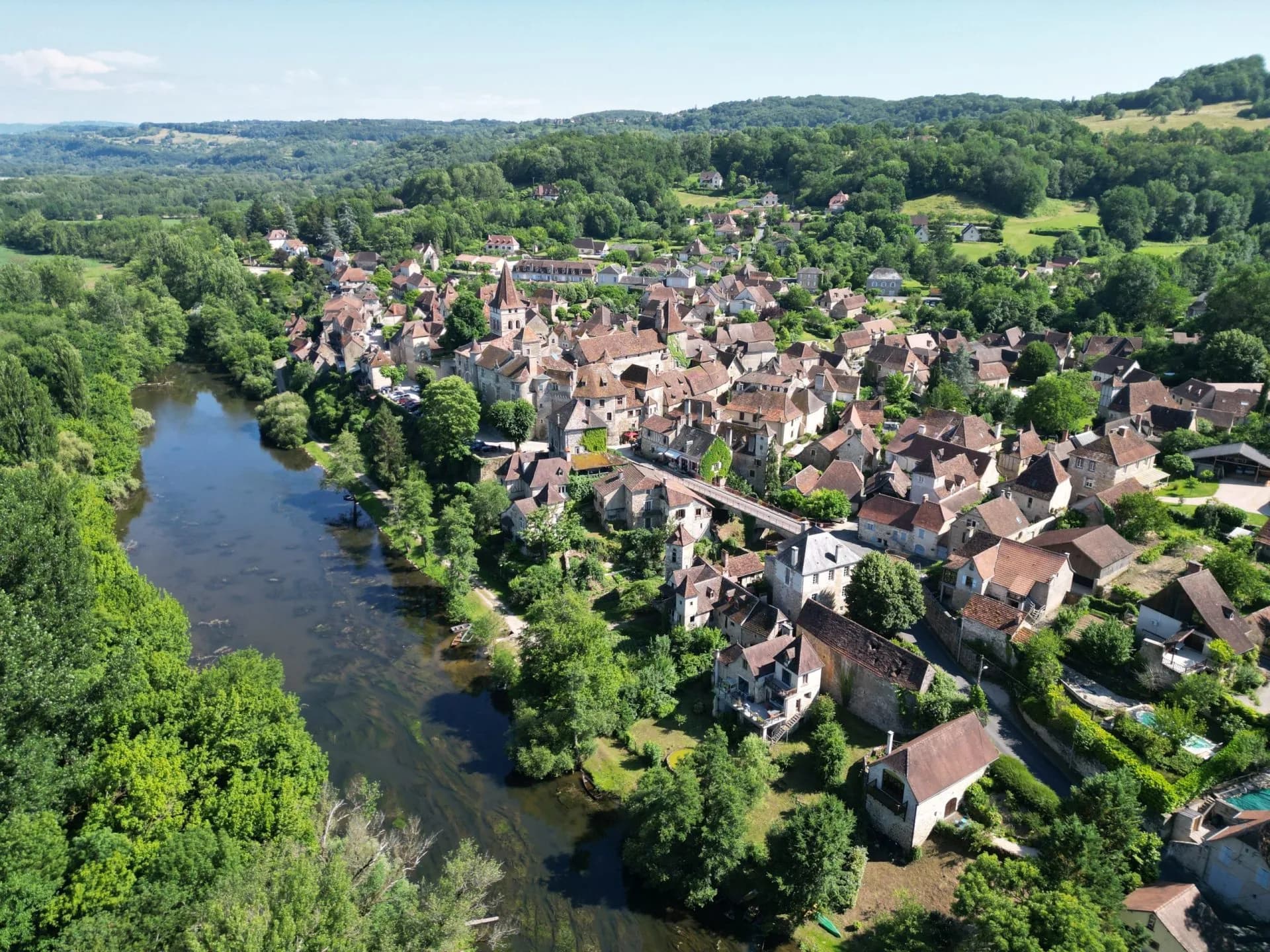 Aerial view of a village with stone houses and red roofs nestled by a river surrounded by lush green forest.
