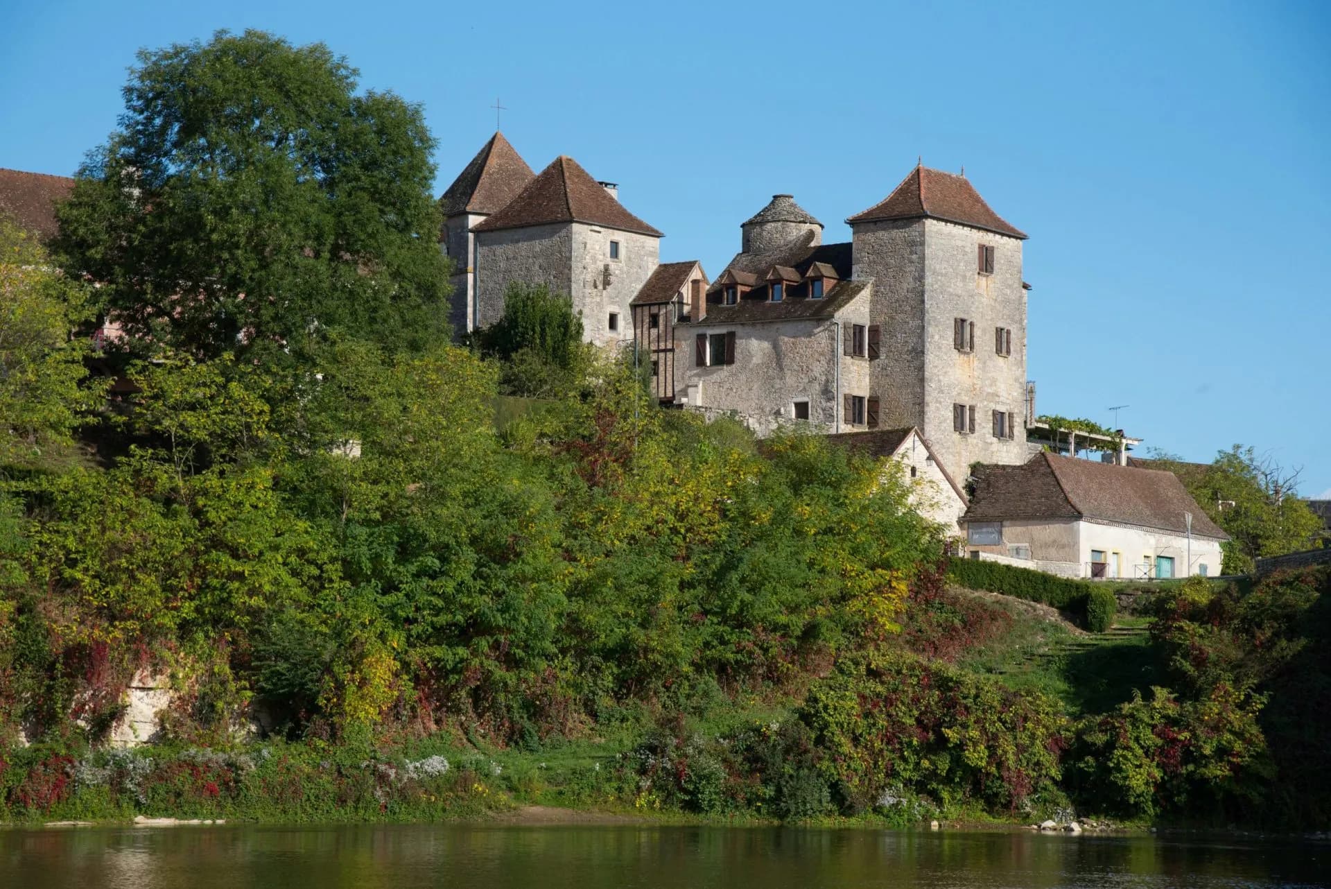 Stone buildings with tiled roofs rise above lush green hillside overlooking calm river water.