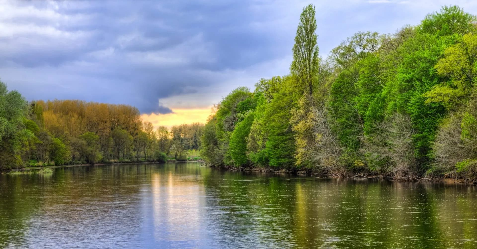 River flowing past lush green and yellow-leaved trees under a dramatic cloudy sky.