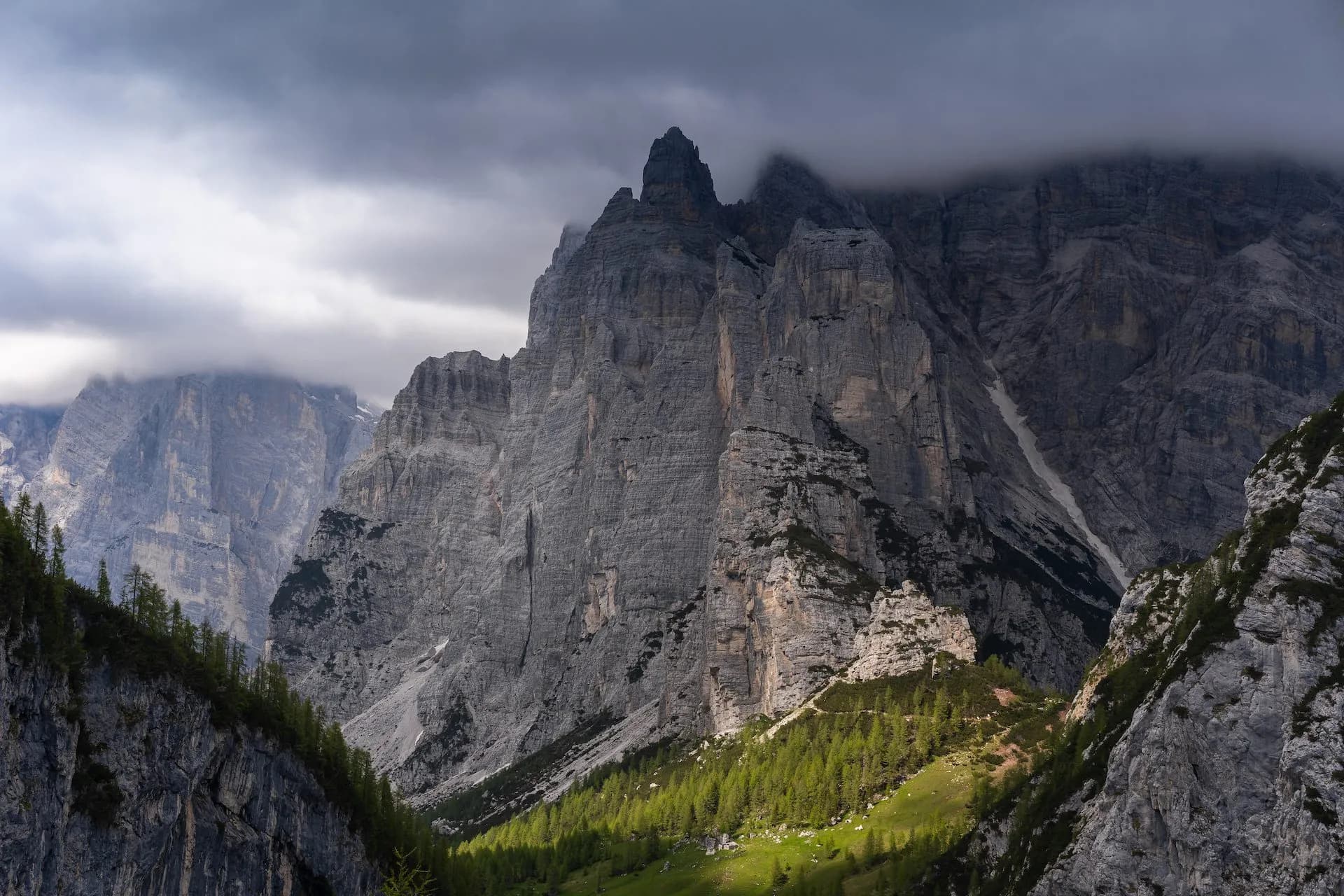 Towering gray mountains under dark clouds with a sunlit green slope and pine trees.