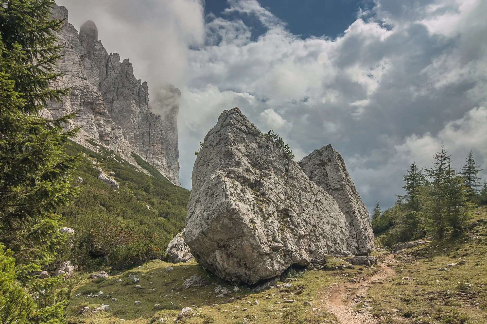 Hiking trail past large boulders near steep, cloudy alpine rock faces with pine trees
