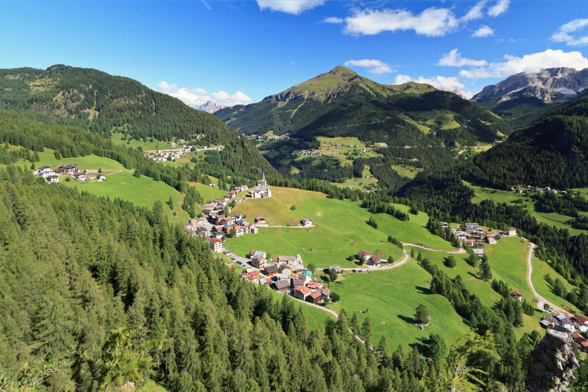 Alpine village nestled in green valley with pine forests and rocky Dolomite peaks under blue sky.