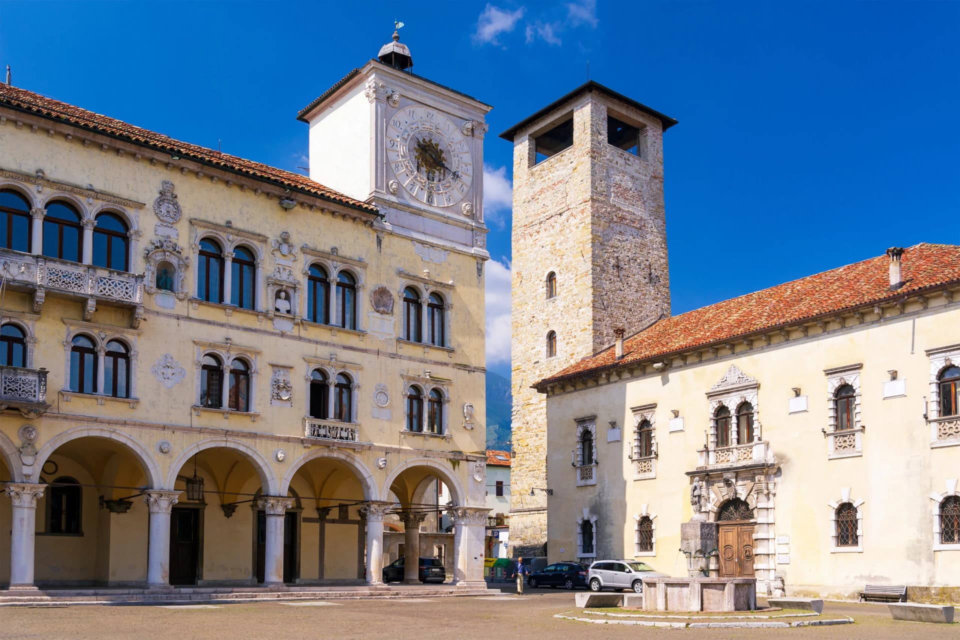 Historic buildings with a clock tower and stone tower in Belluno, Italy under a blue sky.