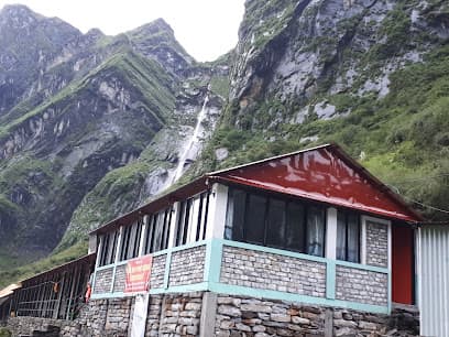 Lodge with stone base and red roof below steep, green mountains and a waterfall at Deurali.