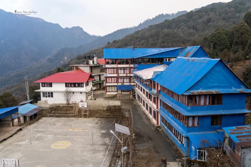 Buildings with blue roofs and a volleyball court set against forested mountains at The Sunny Hotel Ghorepani.