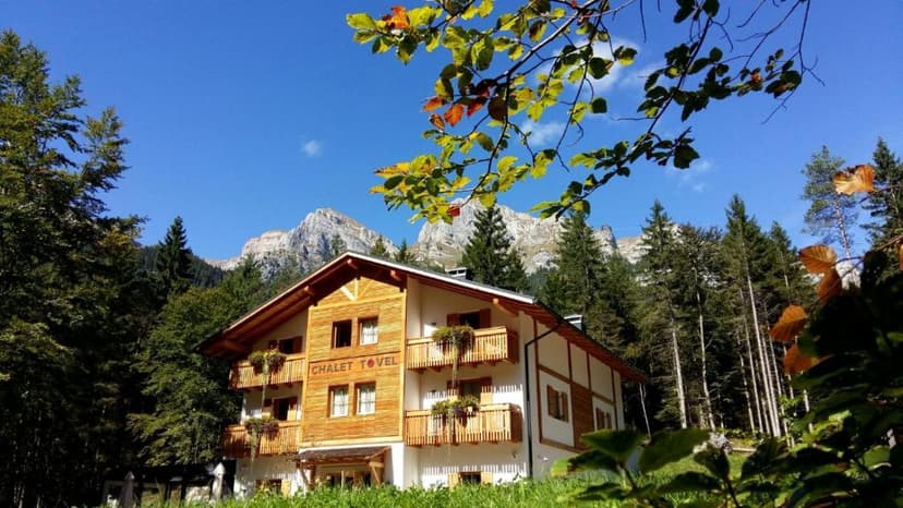 Chalet Tovel with wooden balconies nestled in a forest below rocky mountains under blue sky.