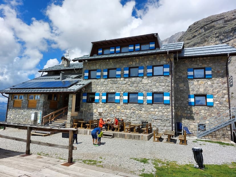 Stone mountain refuge with blue and white shutters, hikers resting outside under cloudy sky.