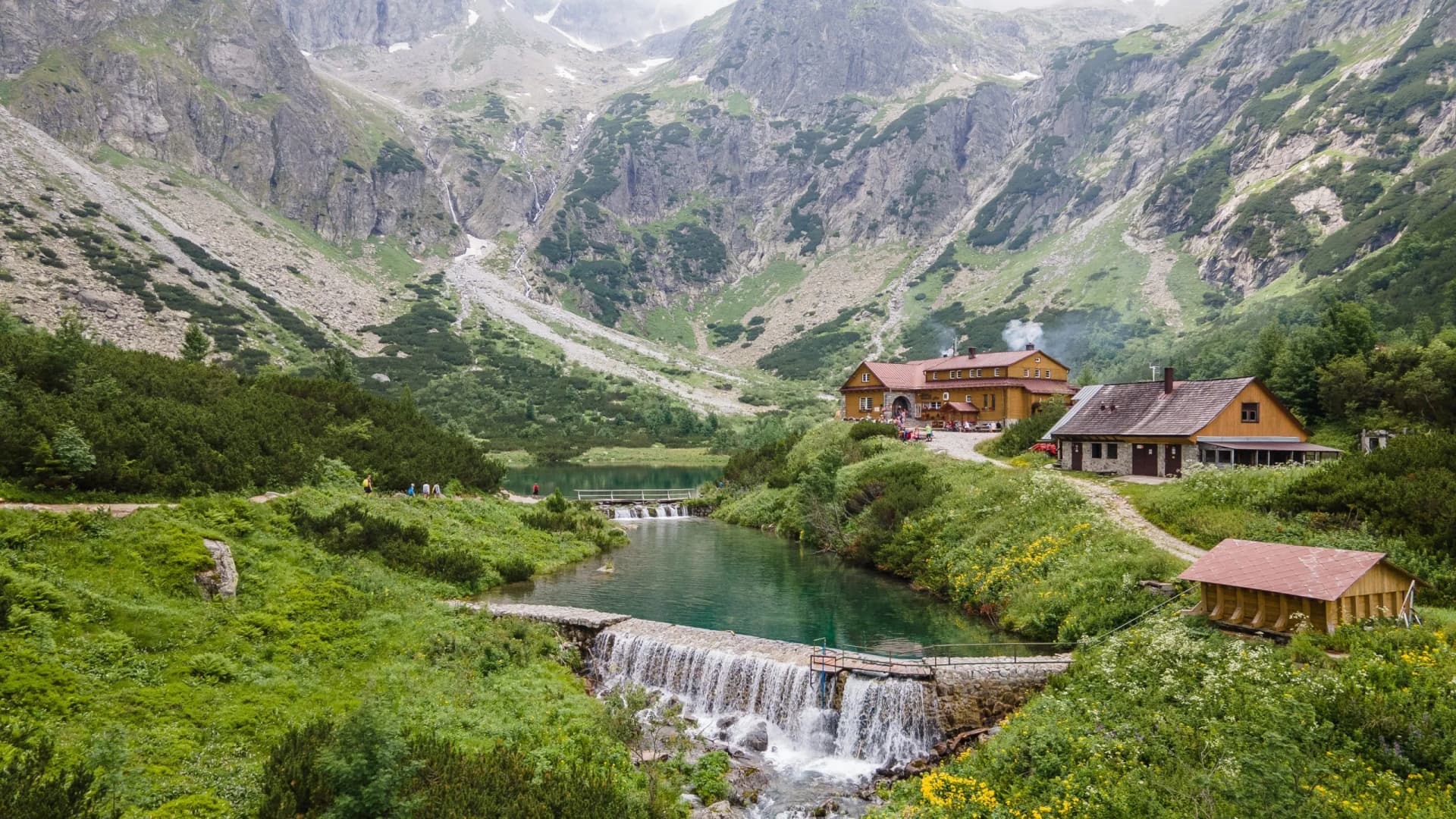 Mountain chalet by green lake with waterfall, hikers near Tatras mountains.