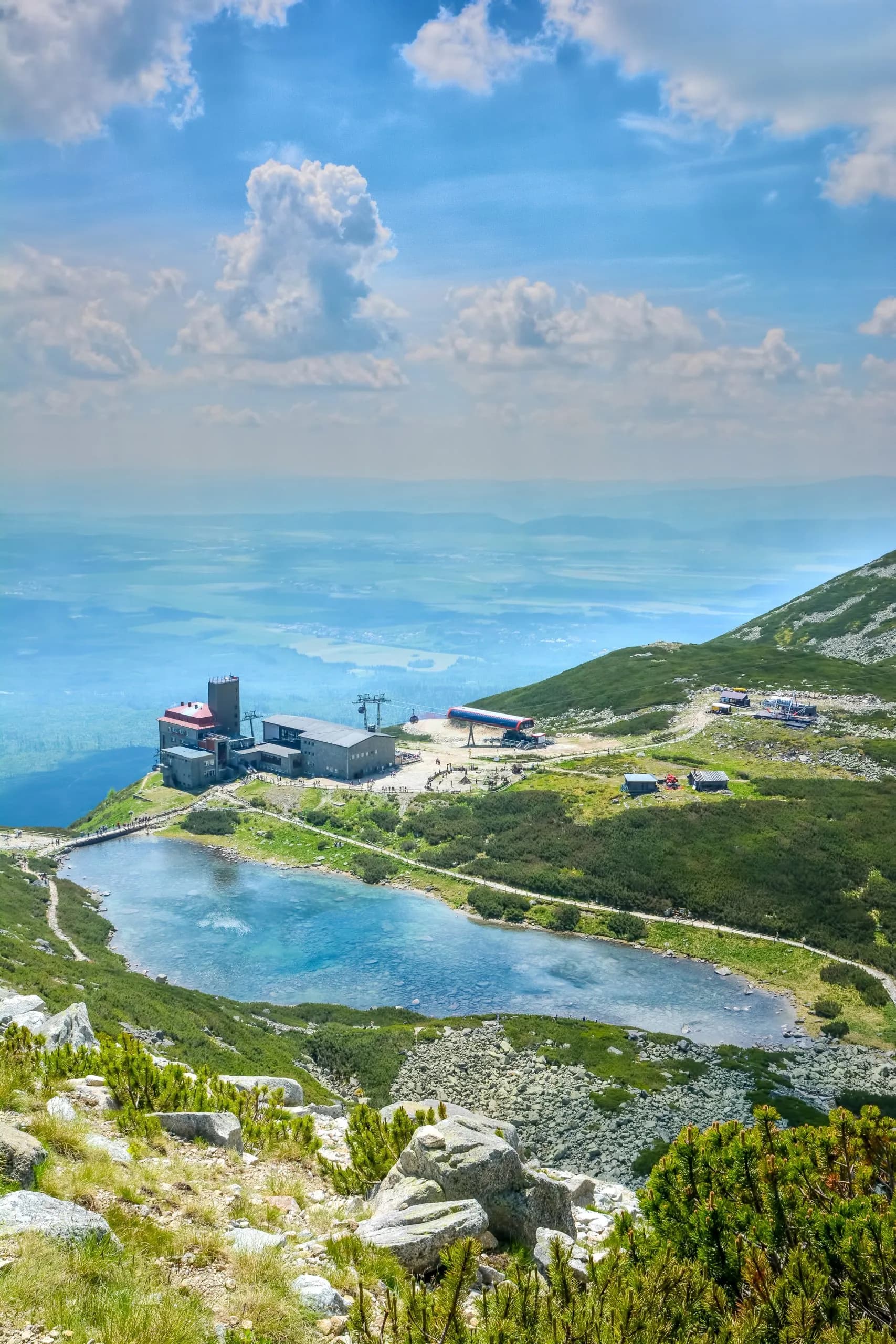 Mountain station buildings beside a blue alpine lake with a vast valley view under a cloudy sky.