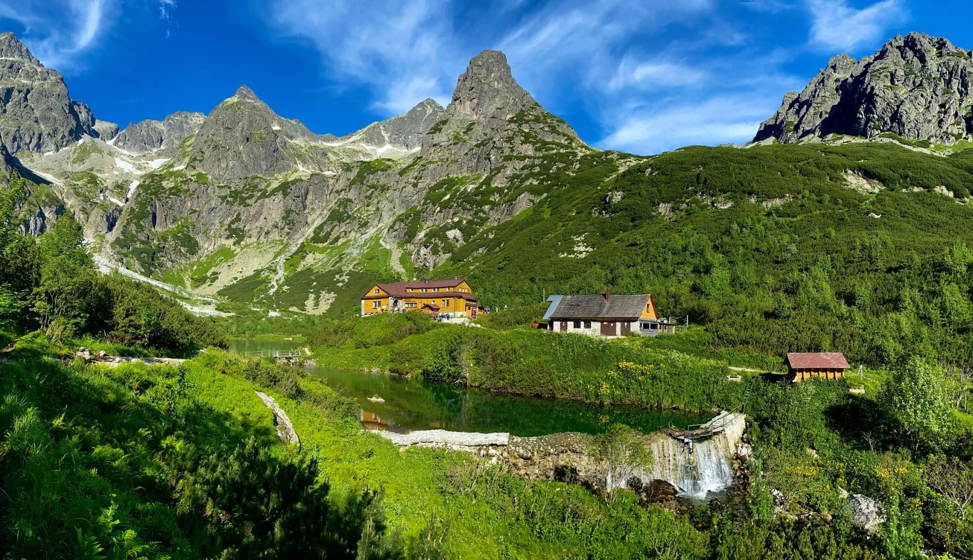 Mountain hut by green lake below rocky peaks with summer foliage, Zelene Pleso.