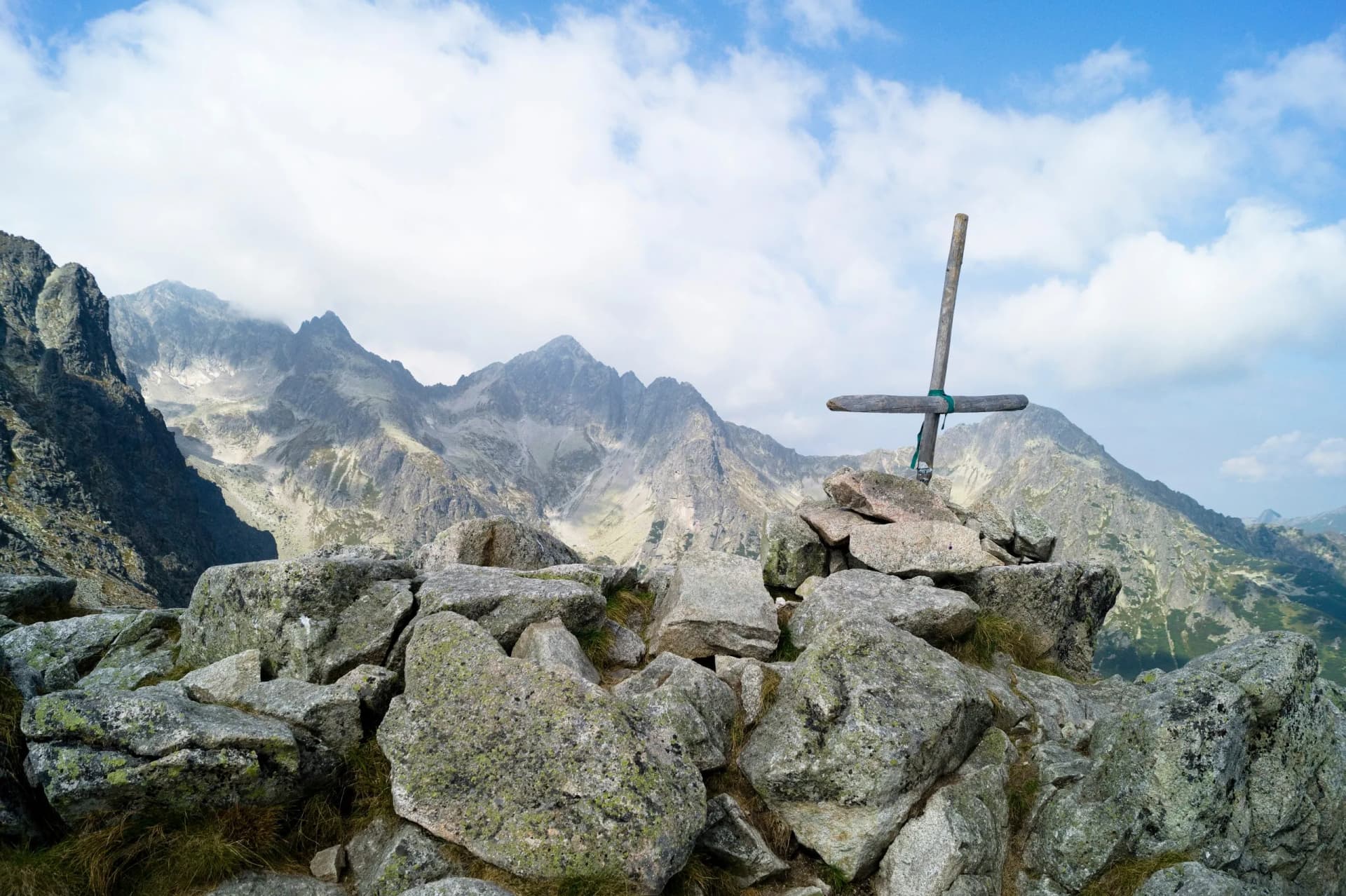 Wooden summit cross on rocks overlooking jagged mountains under a cloudy blue sky.
