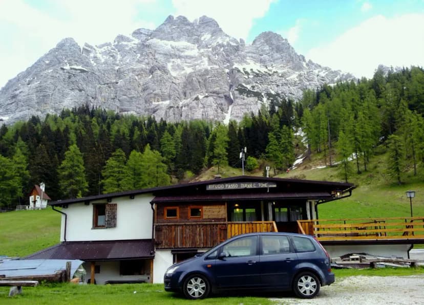 Rifugio Passo Duran C. Tome with car parked in front of snowy mountains and green forest.