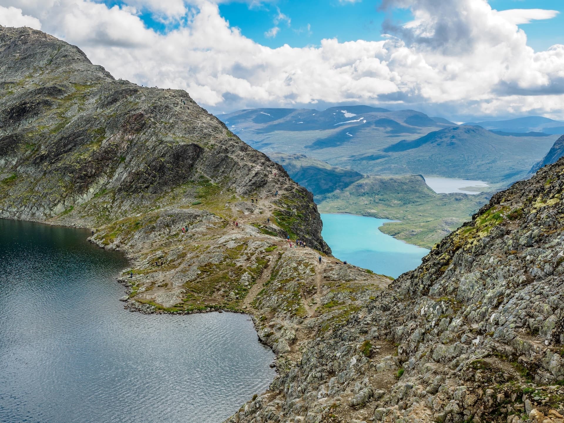 Hikers on rocky Besseggen Ridge trail between dark and turquoise lakes with distant mountains.