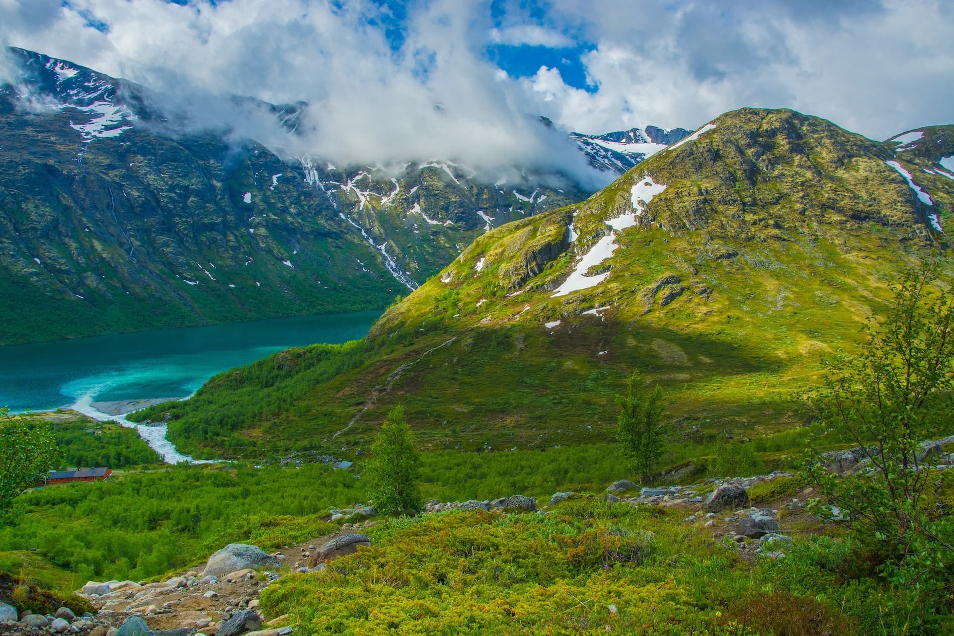 Green mountains with snow patches surround a turquoise glacial lake under a cloudy sky.