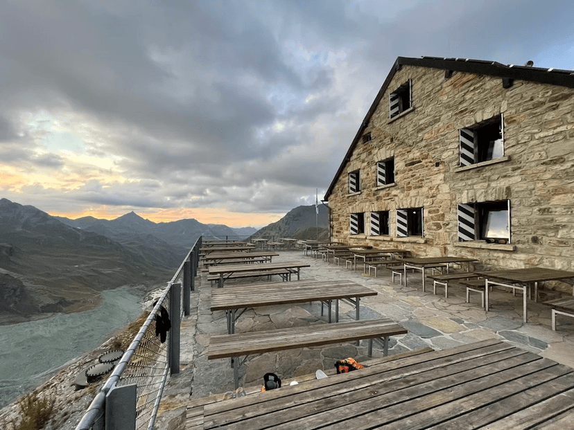 Stone mountain hut terrace with wooden tables overlooking glacial valley and peaks under cloudy sky.