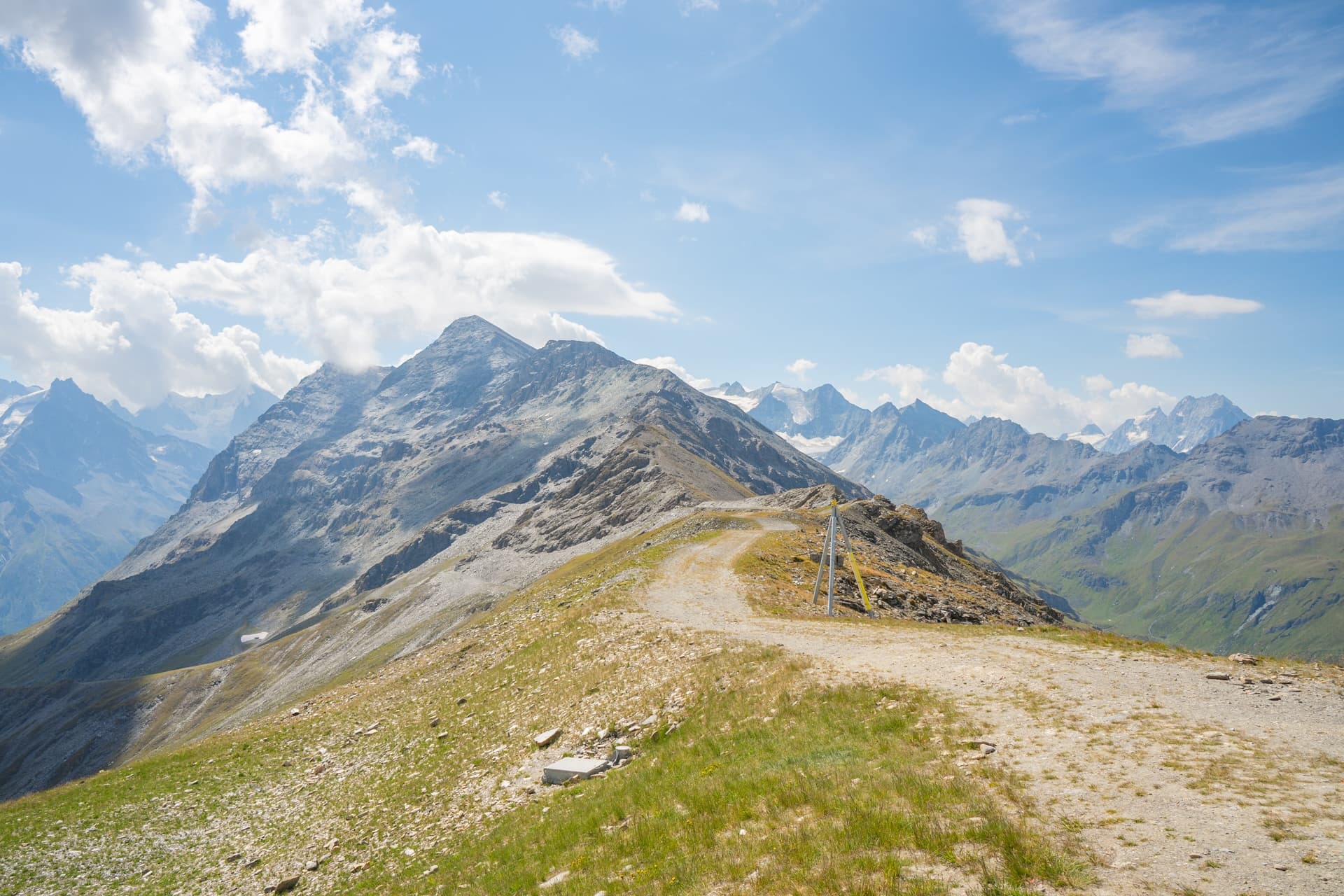 Dirt trail leading up to rocky alpine peaks under a blue sky with white clouds near Lac de Moiry.