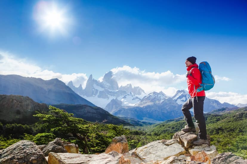 Hiker with backpack overlooking snowy Fitz Roy mountains in Patagonia under bright sun.