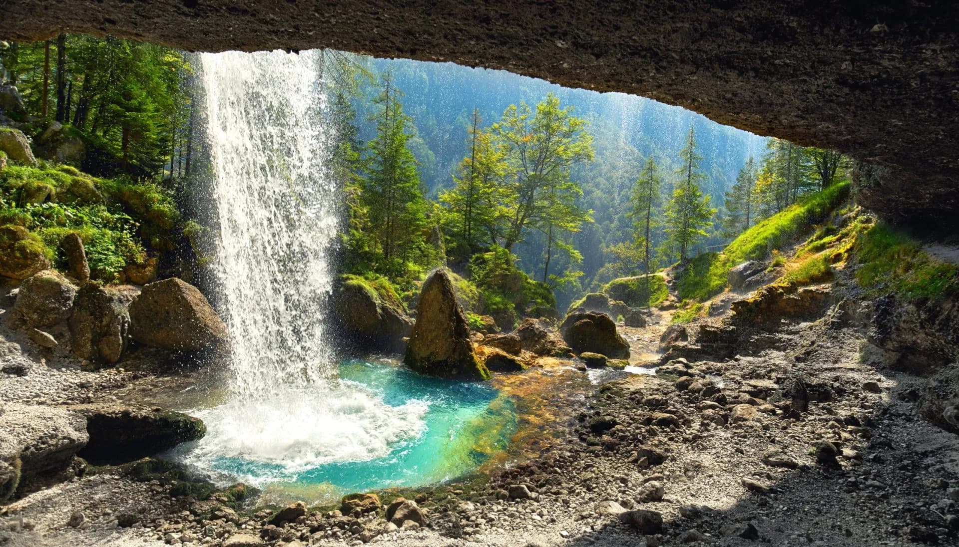 Waterfall cascading into turquoise pool viewed from inside a rocky overhang with forest background.