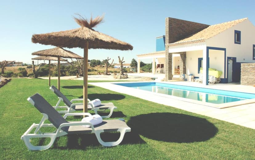 Lounge chairs and thatched umbrellas beside a rectangular swimming pool next to a white villa.