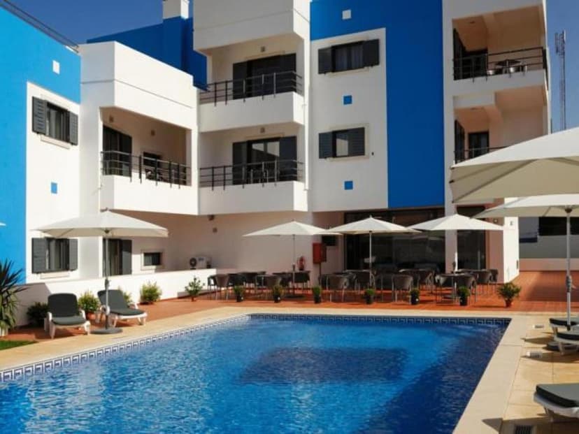 Swimming pool area with white and blue hotel building, lounge chairs, and patio umbrellas.