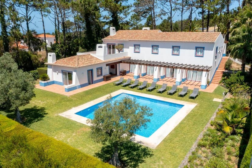 White villa with terracotta roof, swimming pool, and sun loungers surrounded by pine trees.