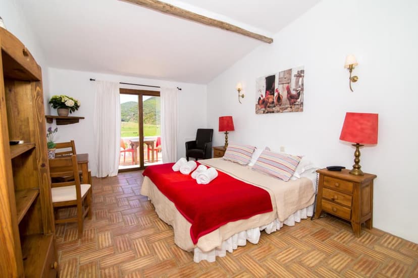 Bedroom with terracotta floors, red bedding, and view of green hills through glass doors.