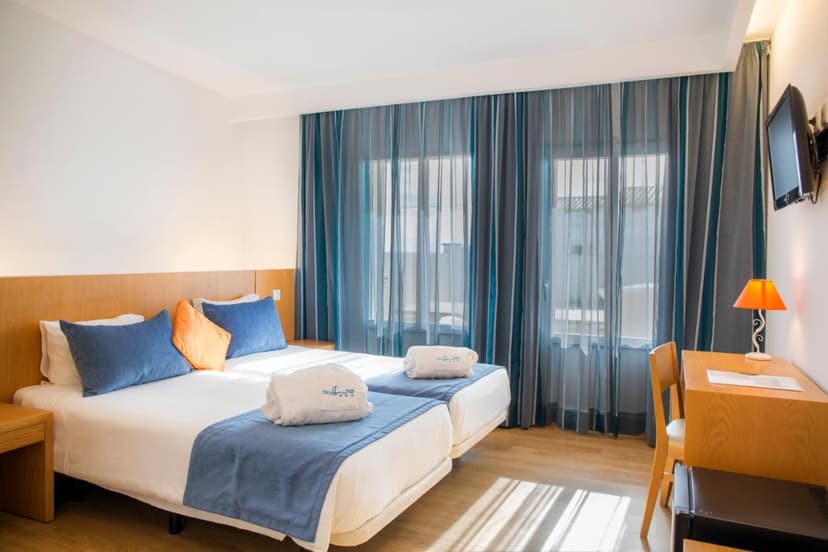Twin hotel room with white bedding, blue accents, wooden furniture, and sheer blue curtains.