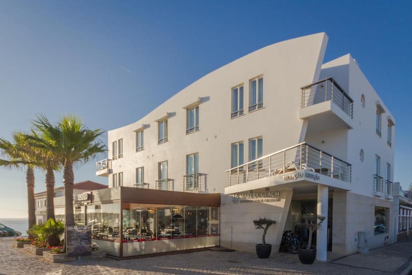 White modern hotel building with "Mareta Beach" sign, palm trees, and cafe under clear blue sky.
