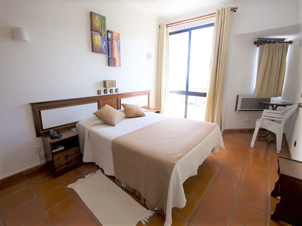 Hotel bedroom with white walls, terracotta tile floor, and a large window with bright light.
