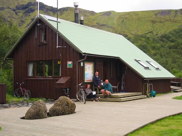 Wooden mountain hut with green roof, people resting outside, and bicycles near grassy hills
