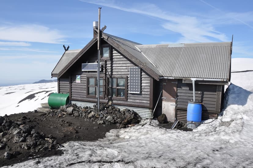 Fimmvörðuháls hut on volcanic ground surrounded by melting snow under a blue sky.
