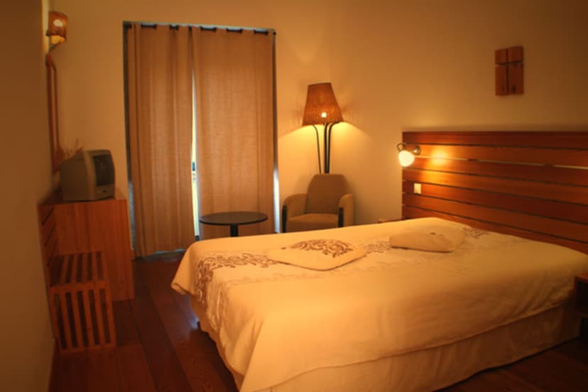 Double bedroom with large bed, wooden headboard, armchair, and window with beige curtains.