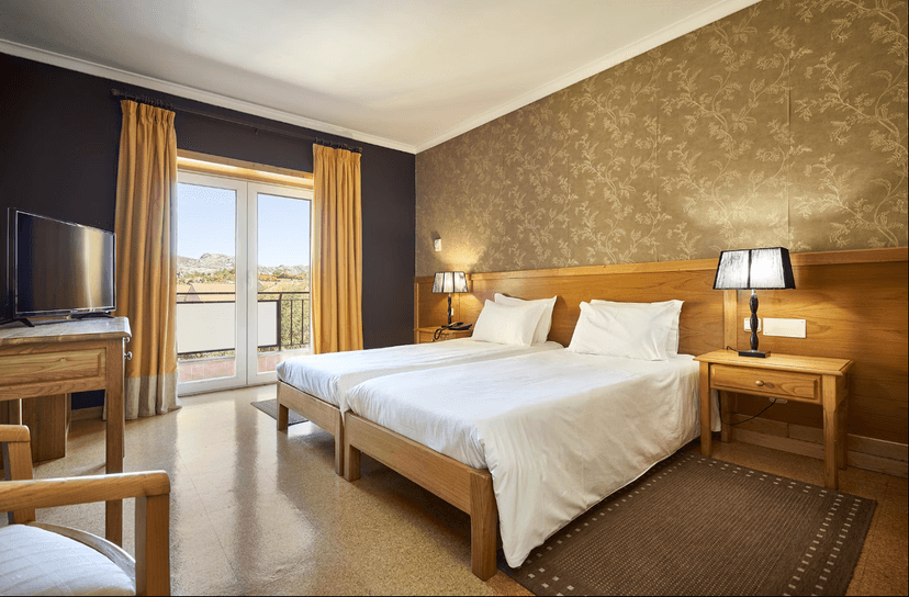 Twin beds in Hotel Castrum Villae room with balcony view of dry, rocky hills.