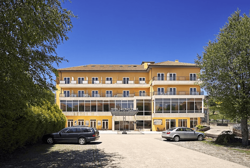 Hotel Castrum Villae exterior: yellow building with balconies under a clear blue sky, cars parked outside.