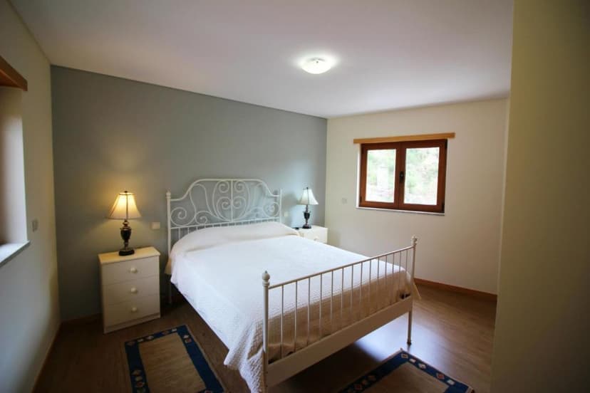 Bedroom with white metal frame bed, nightstands, lamps, and view of greenery through a window.
