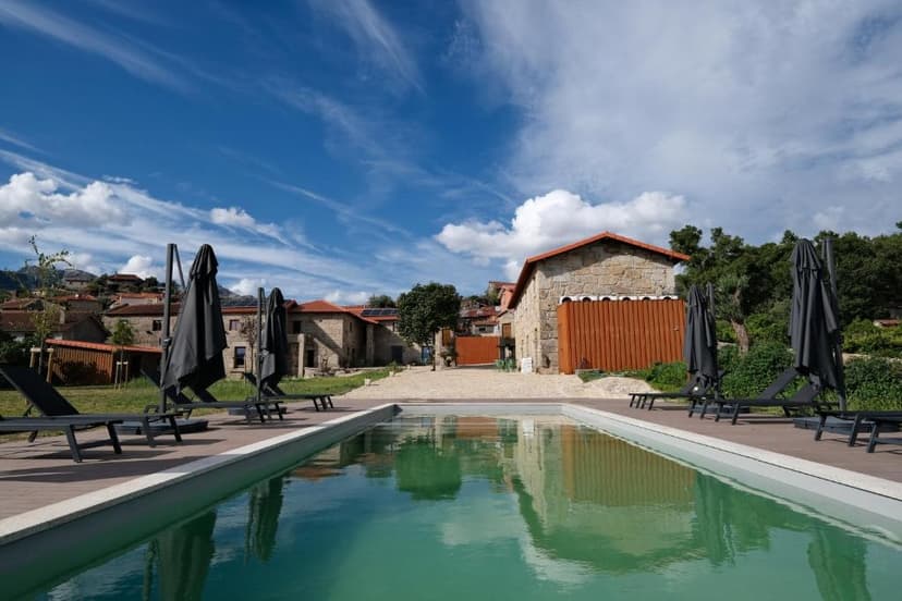 Swimming pool with lounge chairs and folded umbrellas outside stone guesthouse buildings under blue sky.