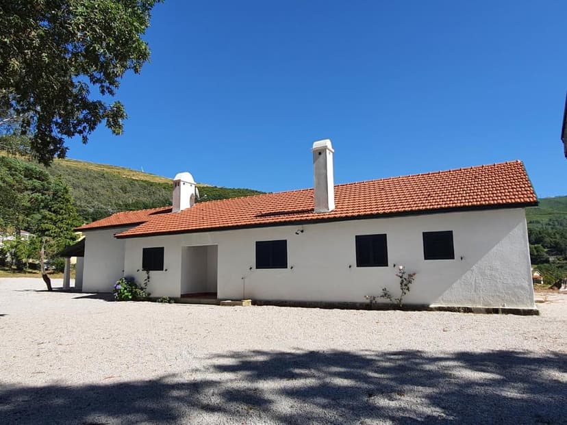 White building with terracotta roof and chimneys in front of a green, grassy mountain under blue sky.