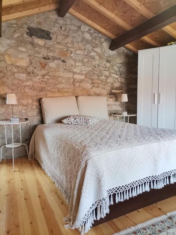 Bedroom with stone wall, sloped wooden ceiling, and bed with white fringed coverlet in Casa da Portela.