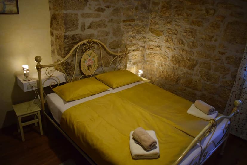 Bedroom with wrought iron bed, yellow linens, and exposed rustic stone wall, Ritossa apartments.