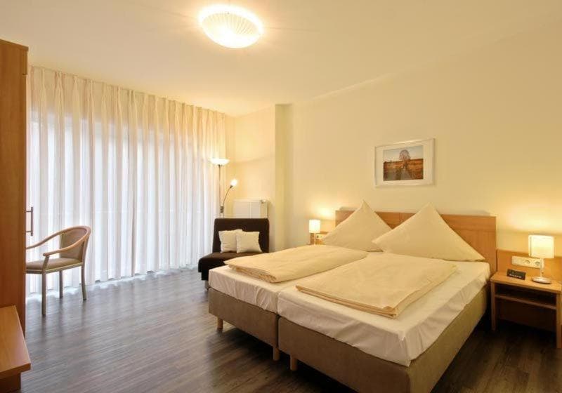 Twin beds with beige bedding in a bright hotel room with sheer curtains and dark wood floor.