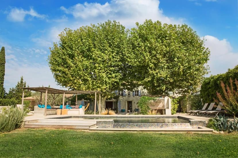Swimming pool area with lounge seating under a pergola near a stone building, shaded by large plane trees.