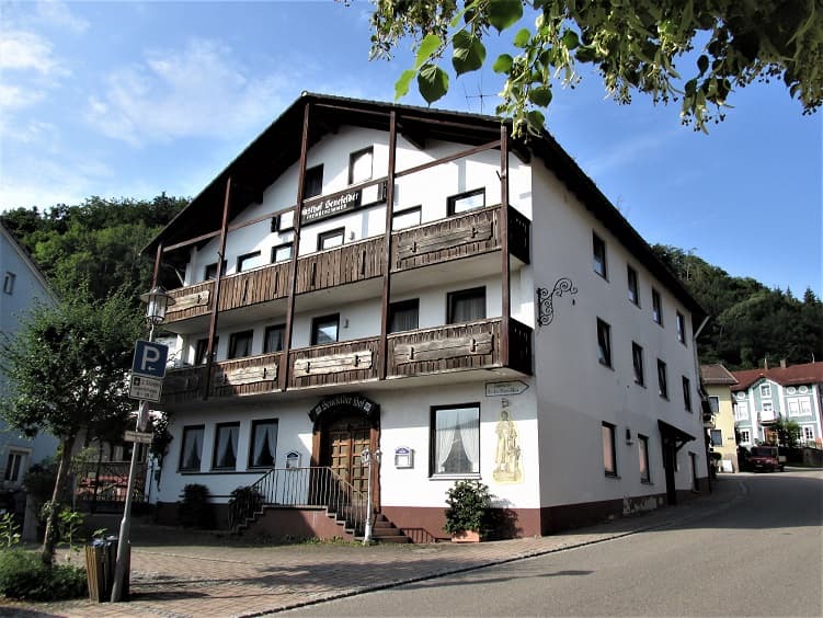 Gasthof Senefelder Hof building with wooden balconies against a backdrop of green hills under a blue sky.