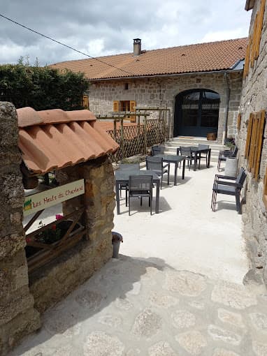 Outdoor seating area with stone buildings and a sign for Les Hauts du Cheylard.