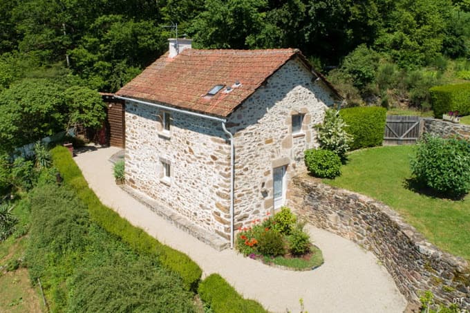 Stone gite with red tile roof surrounded by lush green forest and manicured hedges.
