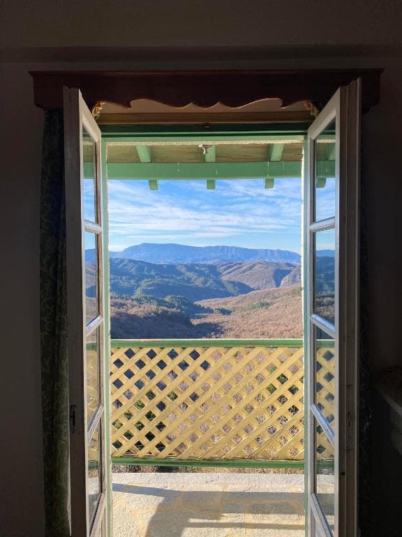 Mountain view from open balcony doors over forested hills under a blue, cloudy sky.